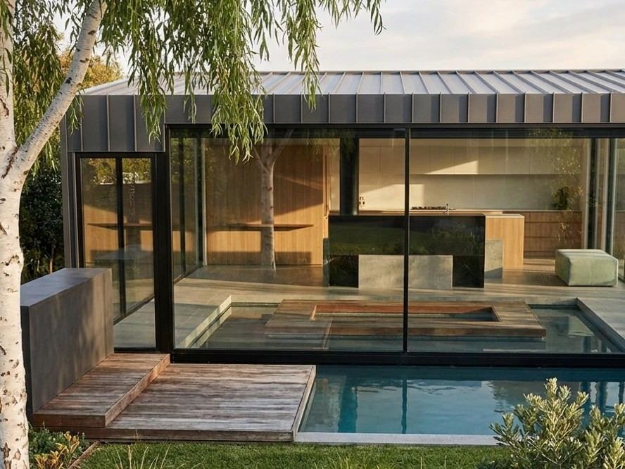 After: the same home, Sonas I, as an AI-enhanced picture: a glass pavilion with a standing-seam roof beside a pool and a silver birch, in late afternoon light. AI-enhanced, indicative.